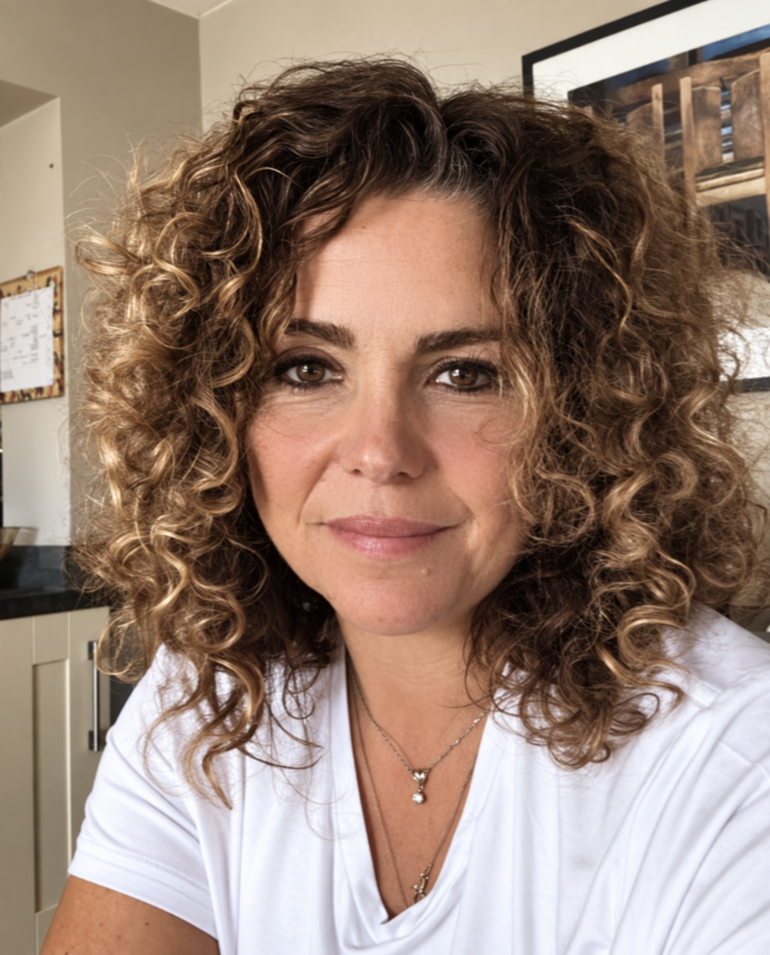 A woman with curly long hair faces the camera, wearing a white top and necklace