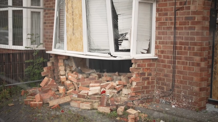 The brickwork under a bay window of a house is smashed and falling on the floor