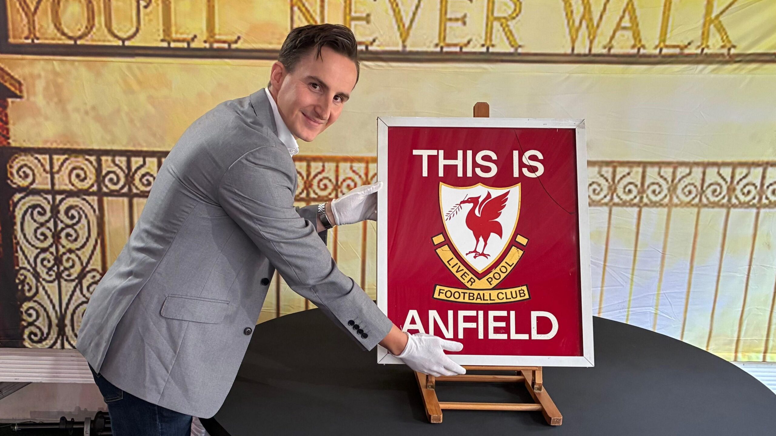 A man in a gray jacket and white gloves stands next to the 'THIS IS ANFIELD' sign and presents it to the camera.