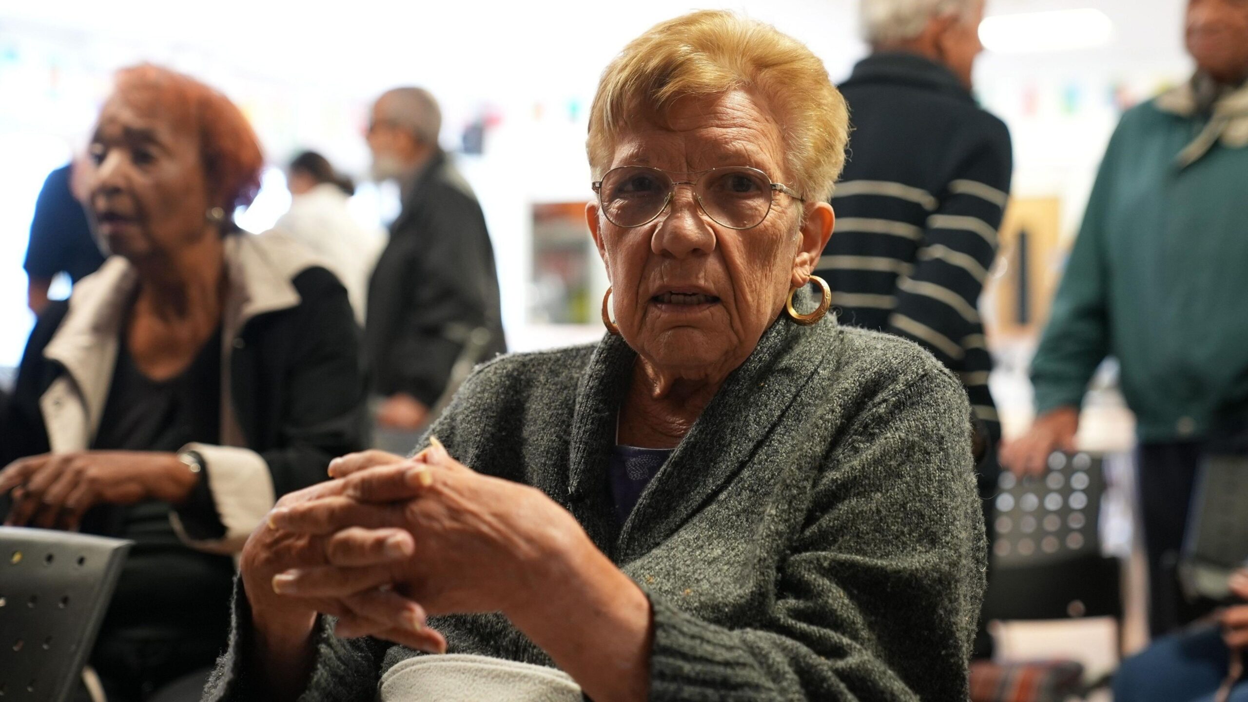 A woman siting in a community centre with her hands clasped - she's wearing a grey cardigan and has dark blonde hair