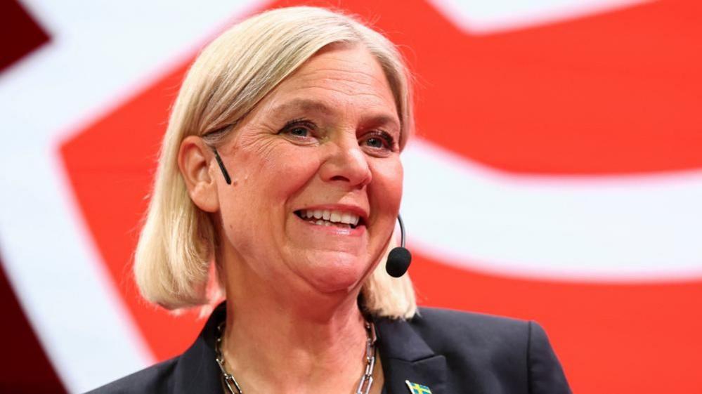 Magdalena Andersson, who has a straight blonde bob and wears a chain silver necklace with a black suit which has a Sweden flag badge on the lapel, speaks into a wearable microphone at a party conference as the general election's voting results are released on Sunday