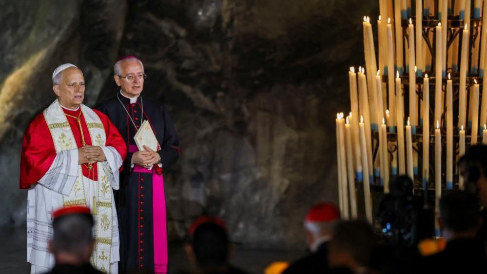 The Pope stands in front of a chandelier of candles as he delivers a prayer. He is wearing red and white decorative rows. A number of men can be seen in front of him and another member of the church stands next to him wearing black and pink robes.