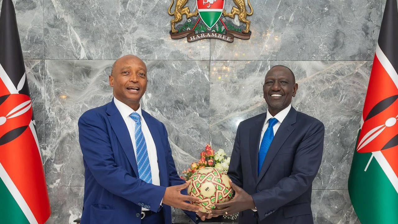 Confederation of African Football president Patrice Motsepe and Kenyan President William Ruto hold a soccer ball between them as they pose for a phot. The two men, both wearing suits, stand in front of a marble wall with a Kenyan coat of arms and a Kenyan flag on either side of the two men