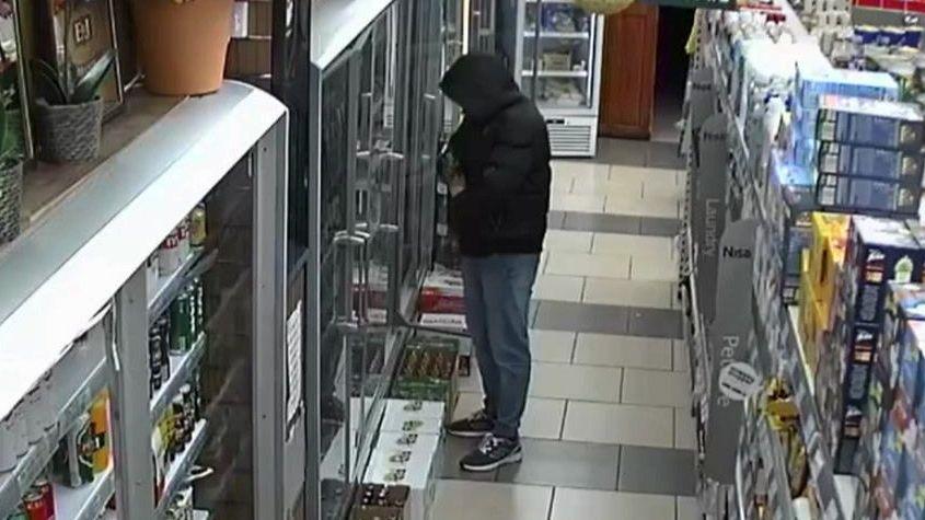 A CCTV image of a man in the aisle of a shop putting bottles in his jacket