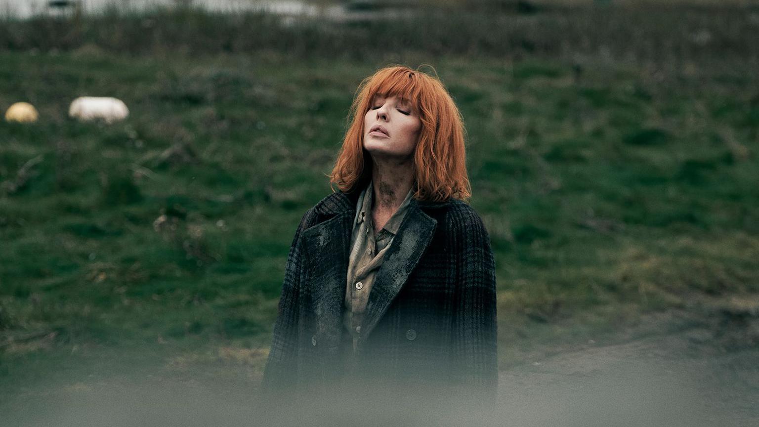 Kelly Reilly standing in a field, looking to the sky with her eyes closed. She has copper shoulder length hair with a fringe. She wears a green shirt and a green tartan coat.