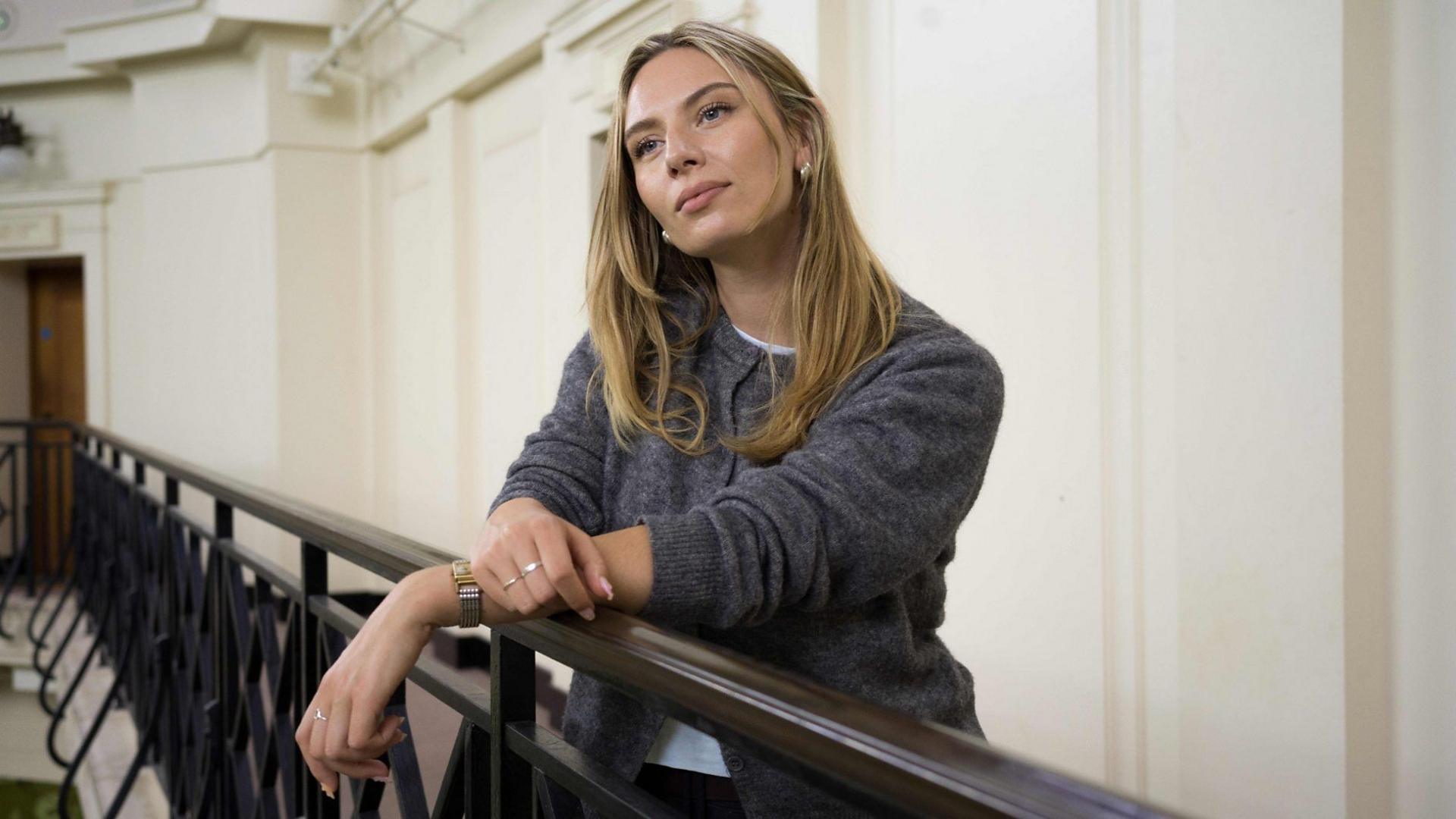 Presenter Sian Eleri is photographed stood next to the rail of a banister. Her right arm is placed over the rail, with her left hand resting on top, just above her wrist. She is wearing a watch, rings and a grey jumper on top of a white t-shirt. She has blonde hair past her shoulders. She is looking away from the camera.