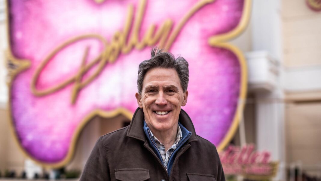 Gavin and Stacey star Rob Brydon’s regret at turning down Dolly Parton interview Rob Brydon smiling at the camera. He had brown hair and wearing a brown jacket. There's a big pink butterfly sign behind him with Dolly written in yellow.