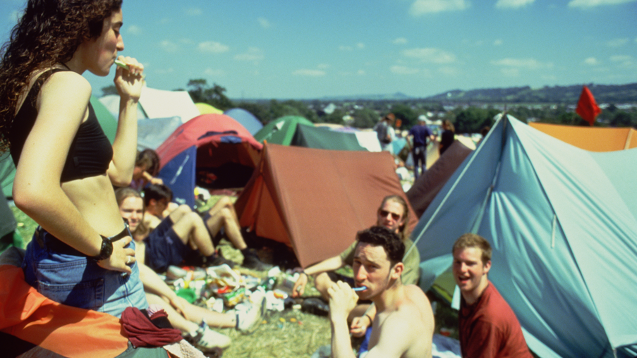 A vintage image of several festivalgoers in a field surrounded by tents - one woman in the foreground is standing up and brushing her teeth and the rest are sitting or lying on the grass, looking to camera. It is a sunny day with a blue sky and a few puffy white clouds