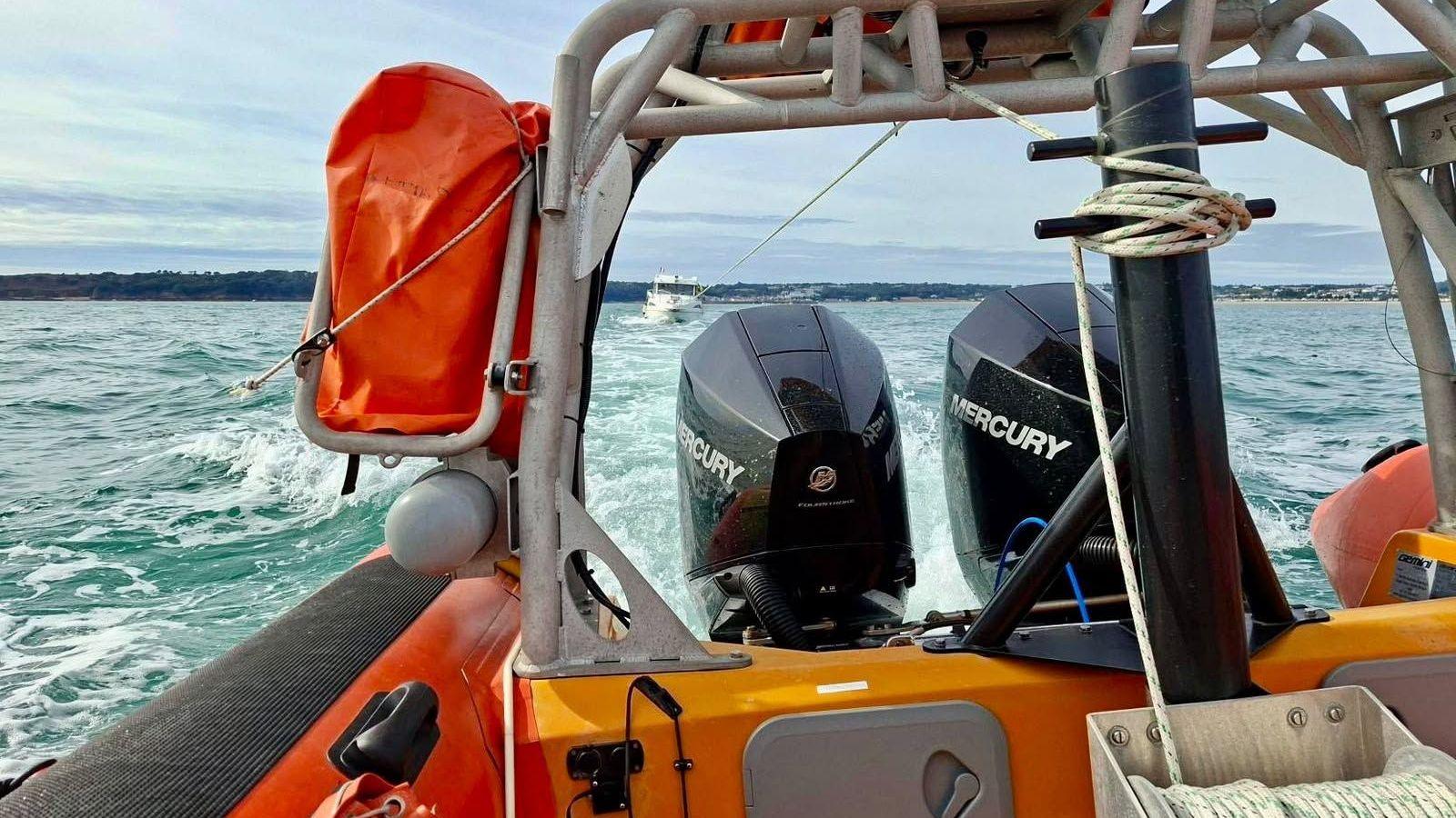 Jersey Lifeboat Association's orange rescue boat travelling across the sea, with two outboard engines visible at the back. Metal framework, ropes and safety equipment are in the foreground, while another boat can be seen in the distance under a cloudy sky.