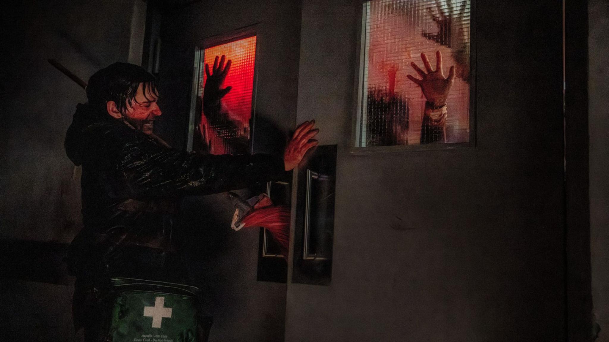 Still from Resident Evil shows Bryan, played by Austin Abrams, desperately pushing against a set of double doors in a hospital hallway. His hands and face are covered in blood, which also matts his hair to his head. A small crowd of zombies can be seen through frosted panes in the doors, their hands pressed against the glass.