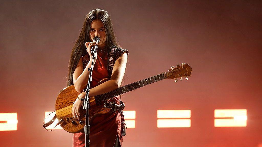 A performer stands center stage holding a guitar and singing into a microphone during a live concert. The performer wears a sleeveless, dark red outfit, while long hair falls over the shoulders.