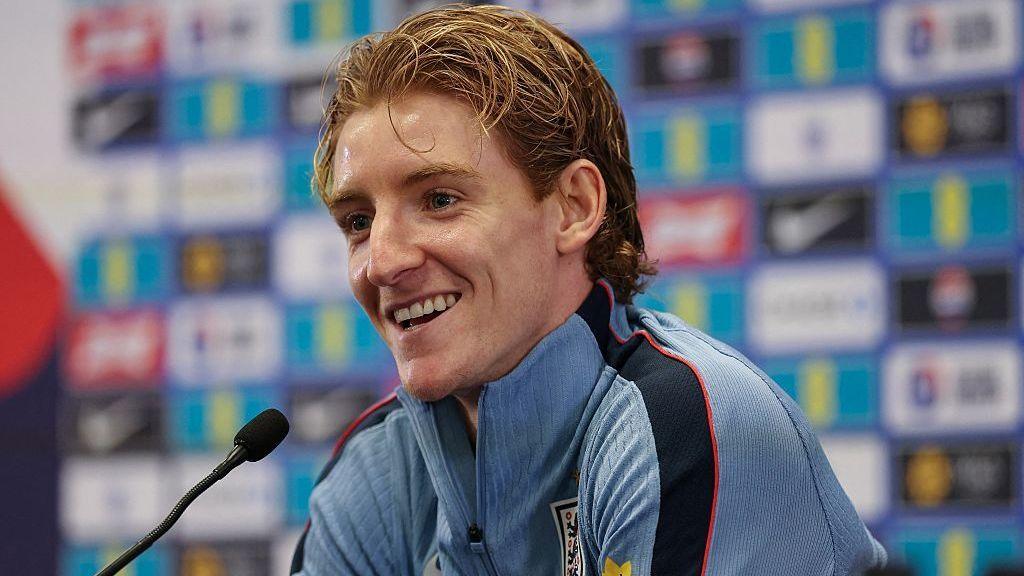A smiling Anthony Gordon, in a blue zip-up top, sits by a microphone at an England news conference