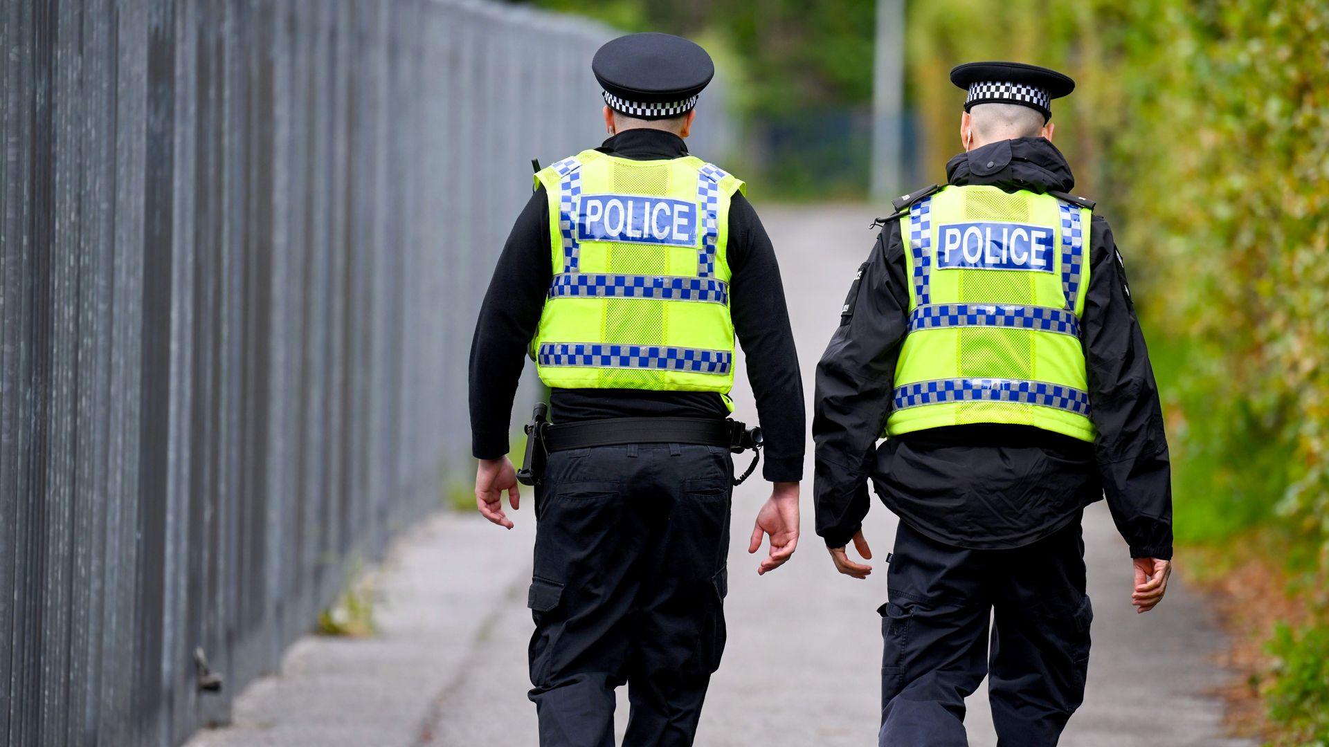 Two police officers are walking down a street. They are both wearing hi-vis vests over a black uniform and wearing black hats. There are bushes to their right and a metal fence to their left.