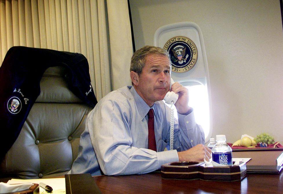 President Bush is at a desk on the plane with his jacket off and hung on the back of his seat, he is wearing a light blue shirt and maroon tie. He has a phone in his left hand and seems to be in conversation.