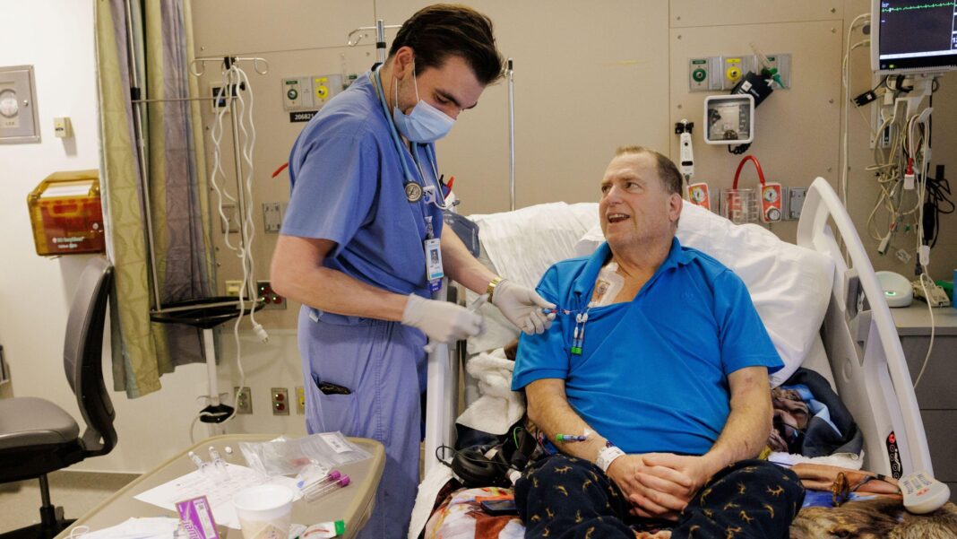 The image shows a hospital room with a patient seated in an adjustable bed, wearing a bright blue polo shirt and patterned dark trousers. A healthcare worker dressed in blue scrubs, gloves, a face mask, and a stethoscope is standing beside the bed and handling tubing connected to a medical access point on the patient's upper chest. Various items including syringes, medical packaging, paperwork, and a disposable cup are arranged on a bedside table in the foreground. The room contains hospital equipment such as wall-mounted oxygen and suction outlets, monitoring devices, intravenous poles, and cables connected to equipment near the bed.