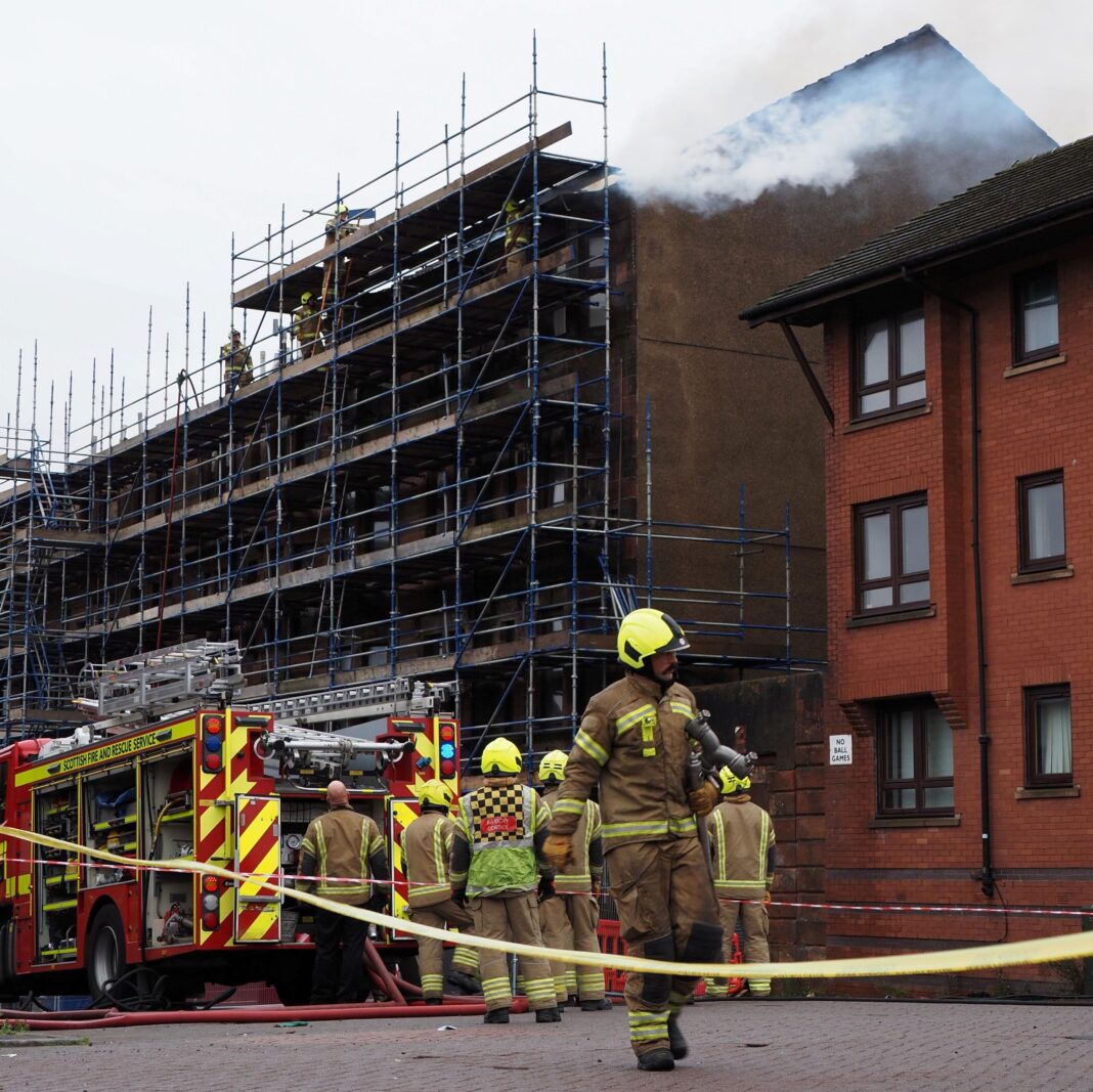 Police probe cause of fire at Glasgow affordable homes tenement Fie engine and a number of firefighters in a cordoned-off area of a road. There is snoke coming from the roof of the building which is covered by scaffolding.