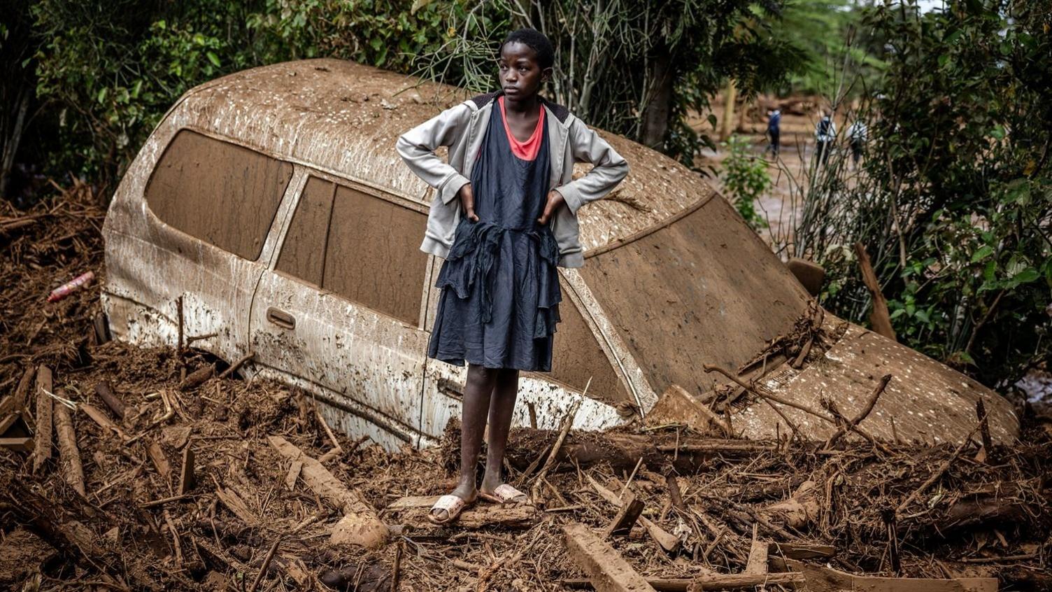 A young girl stands in a blue dress and grey cardigan with sandals, in front of a car buried in a flood amongst debris