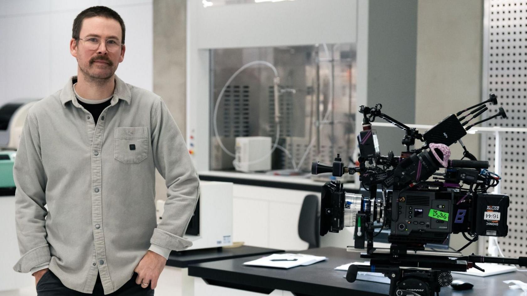 Zach Cregger, pictured on the set of Resident Evil. He's standing in what looks like a lab, with bright white walls and equipment in the background. He's posing next to a large film camera, with various attachments and accessories attached to it.