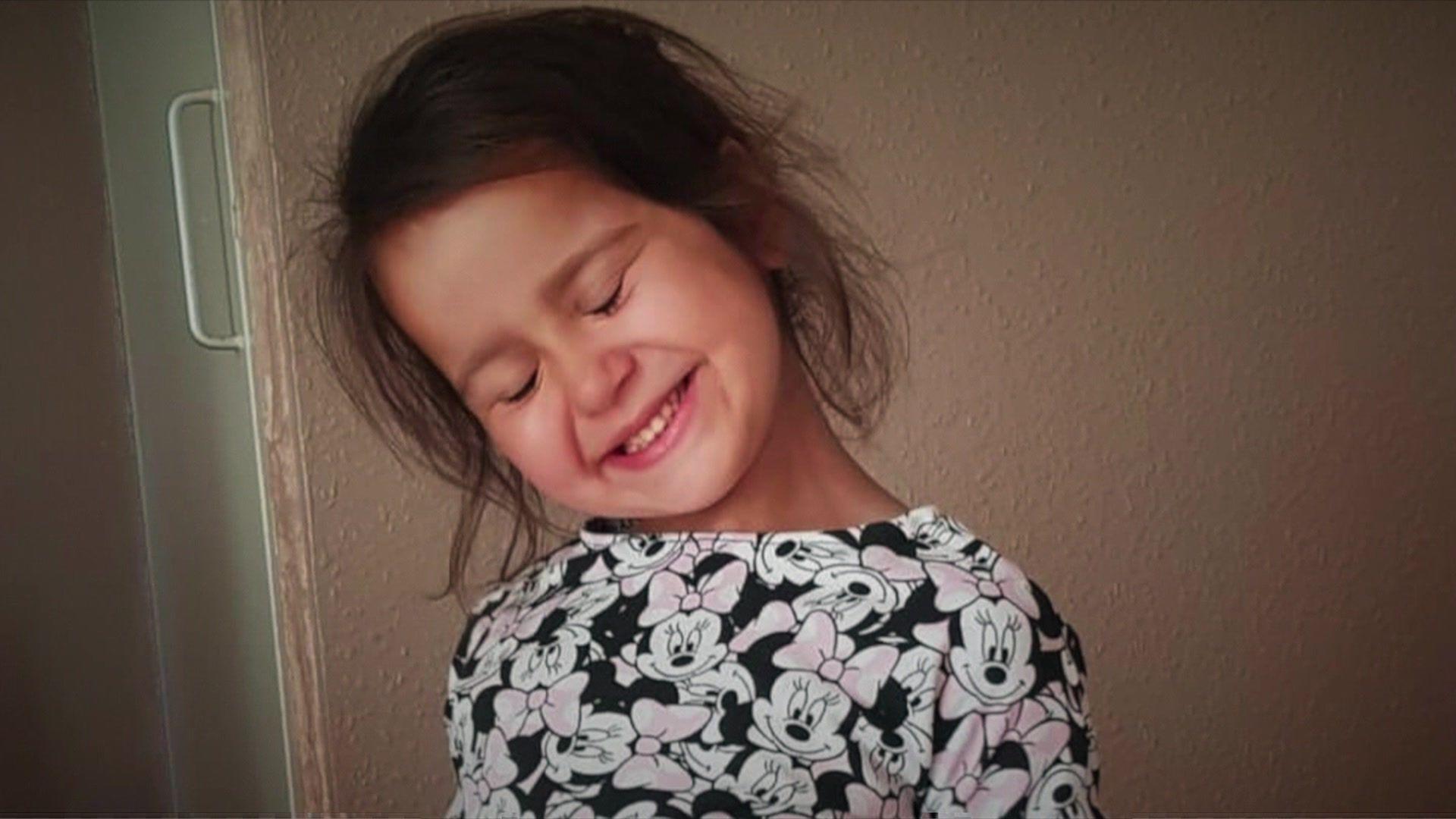 A young girl in a Minnie Mouse dress smiles with her head tilted to the side.