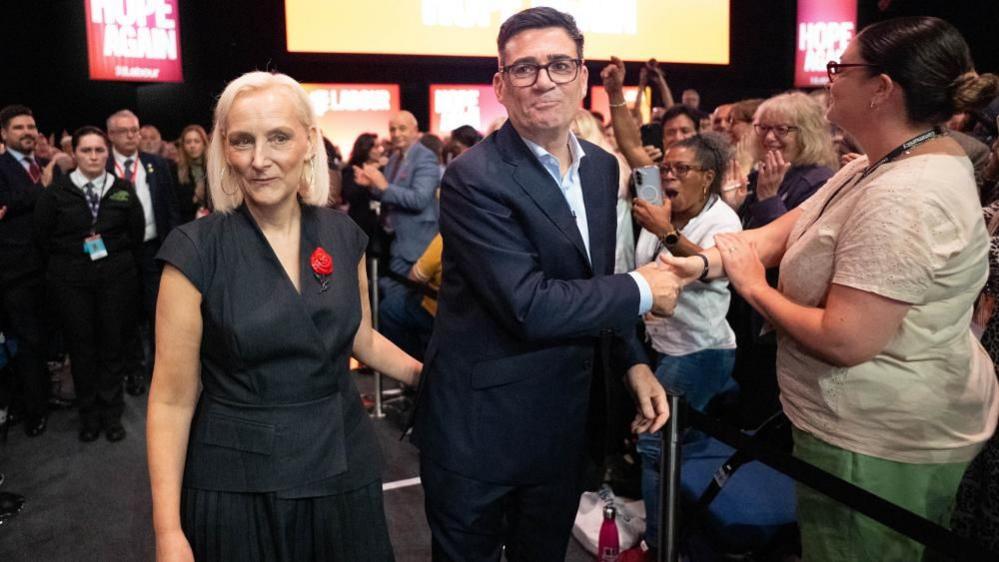 Marie-France van Heel and Andy Burnham greet delegates in the audience after the prime minister's keynote speech. Van Heel is wearing a dark-coloured dress with no sleeves. Burnham is wearing a dark-coloured jacket, white shirt and glasses.