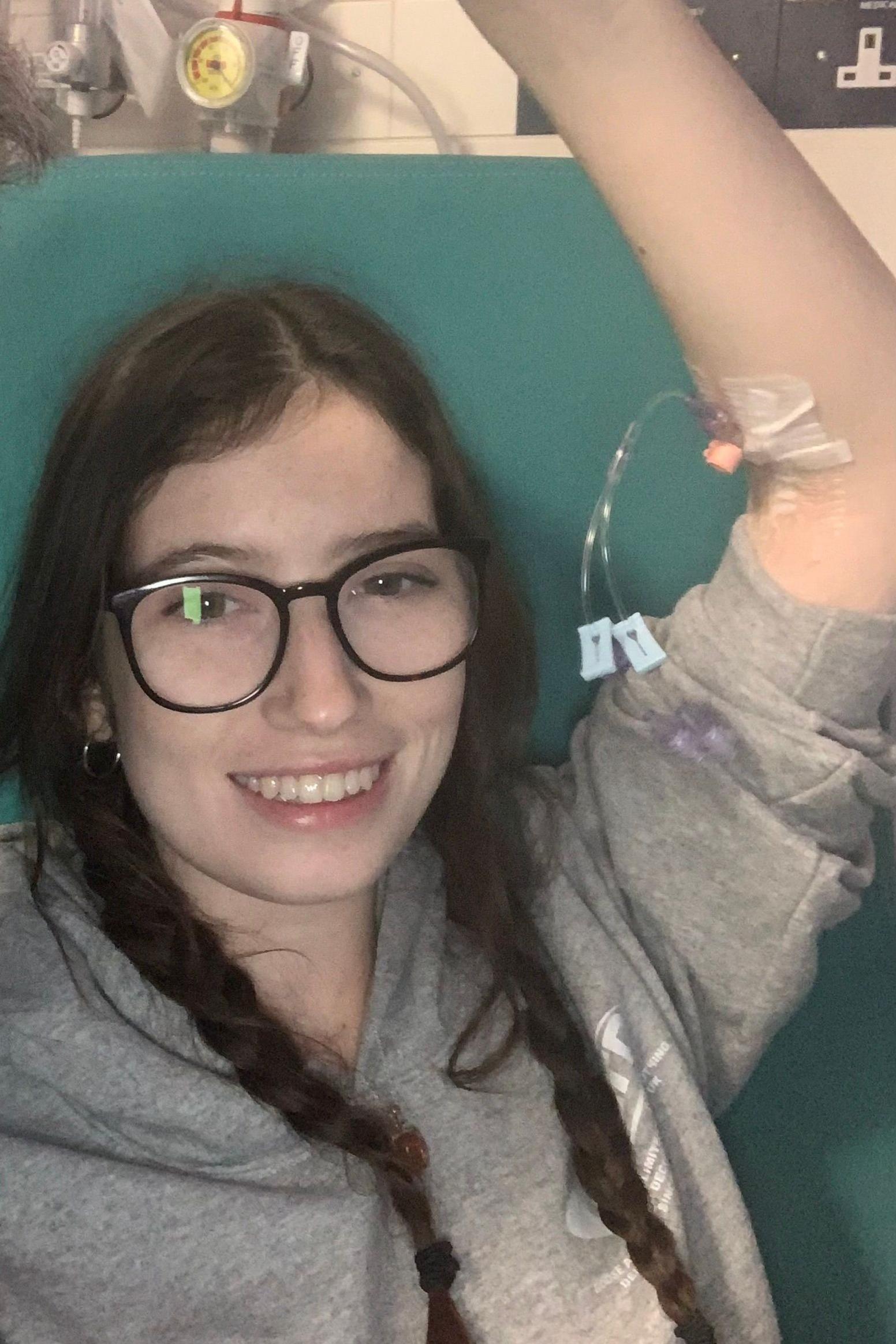 A girl with long dark hair in two plaits. She is wearing a grey hoodie and has one arm raised above her head with an IV needle in it. She is wearing glasses, and is smiling as she looks at the camera.