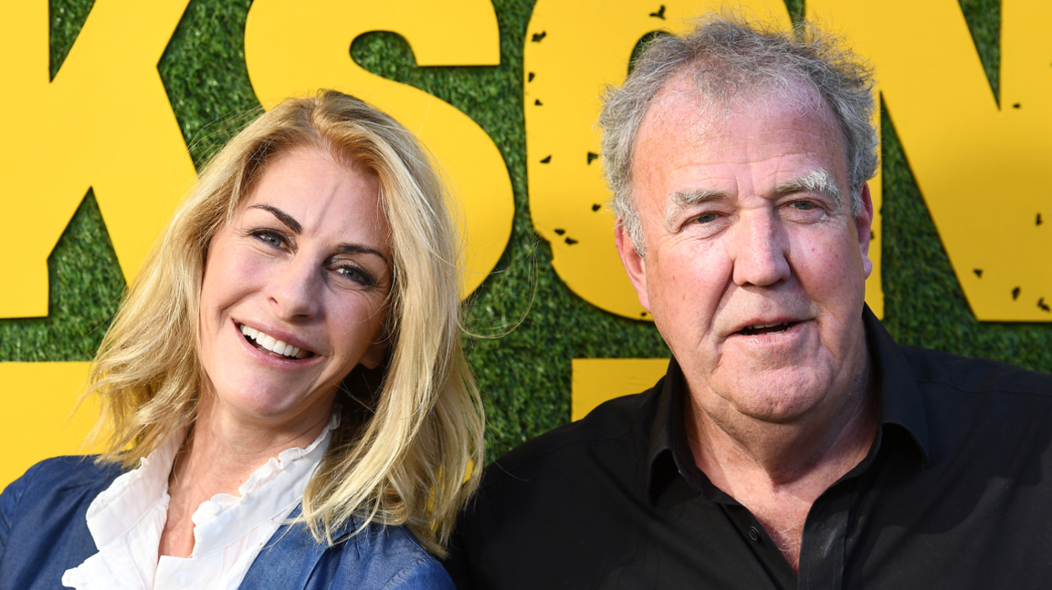 Lisa Hogan and Jeremy Clarkson pose for a head-and-shoulders shot. Lisa has long blonde hair and is wearing a blue jacket over a white shirt. She is smiling widely at the camera. Clarkson, to the right, is wearing a black shirt and looking in the direction of the camera with his mouth slightly ajar,