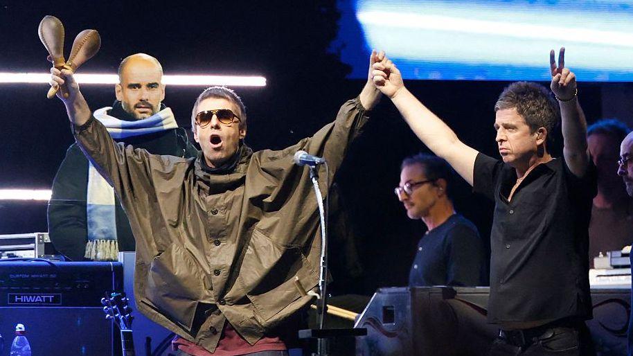 Noel and Liam Gallagher at one of the Oasis reunion gigs. They are holding their arms in the air and touching hands in the middle. Liam is holding maracas in his other hand while Noel is doing a peace sign with his fingers. Other band members are in the background.