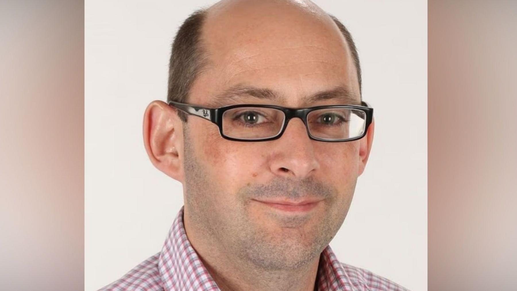 Dr Mark Deakin, who is bald, has glasses, and is wearing a red and white check shirt.