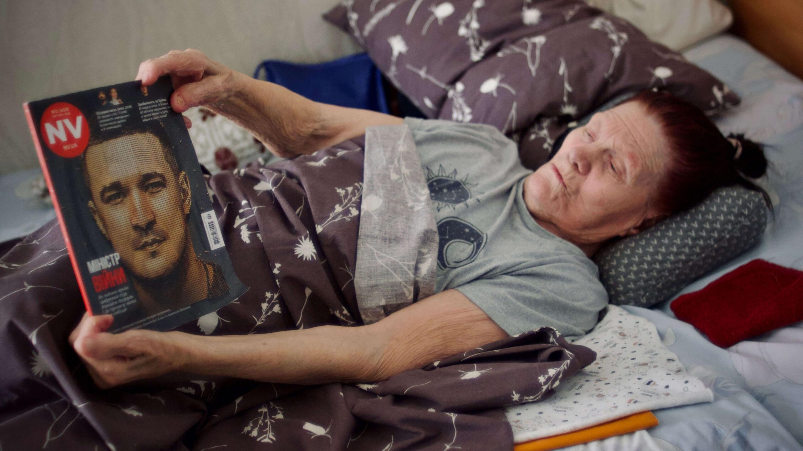 Nadia is pictured in bed reading a magazine. She's wearing a grey t-shirt and has dark hair