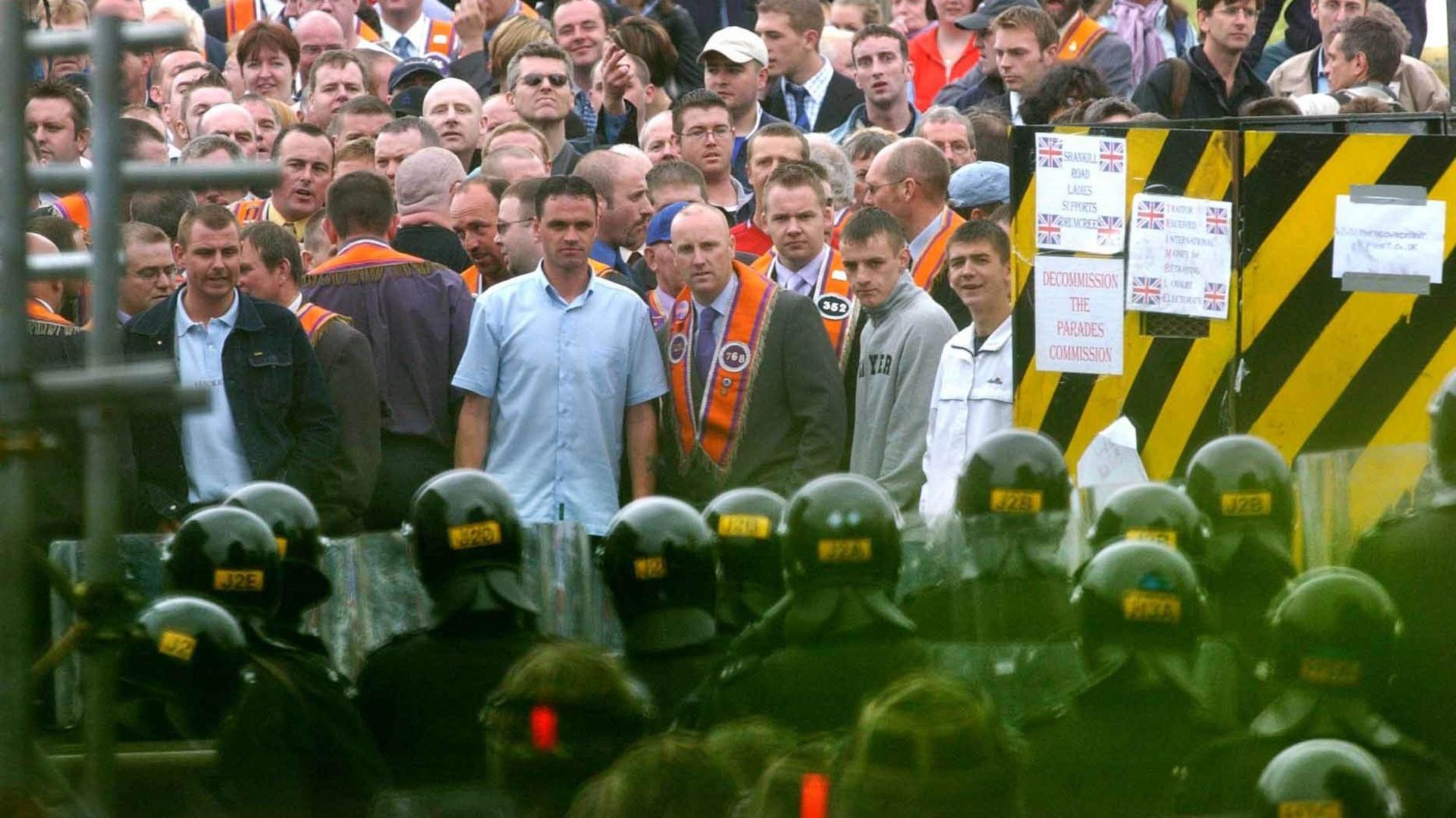 A group of police in riot gear are facing a large crowd of people. Mostly are men. Many are wearing orange sashes. Signs say decommission the parades commission.