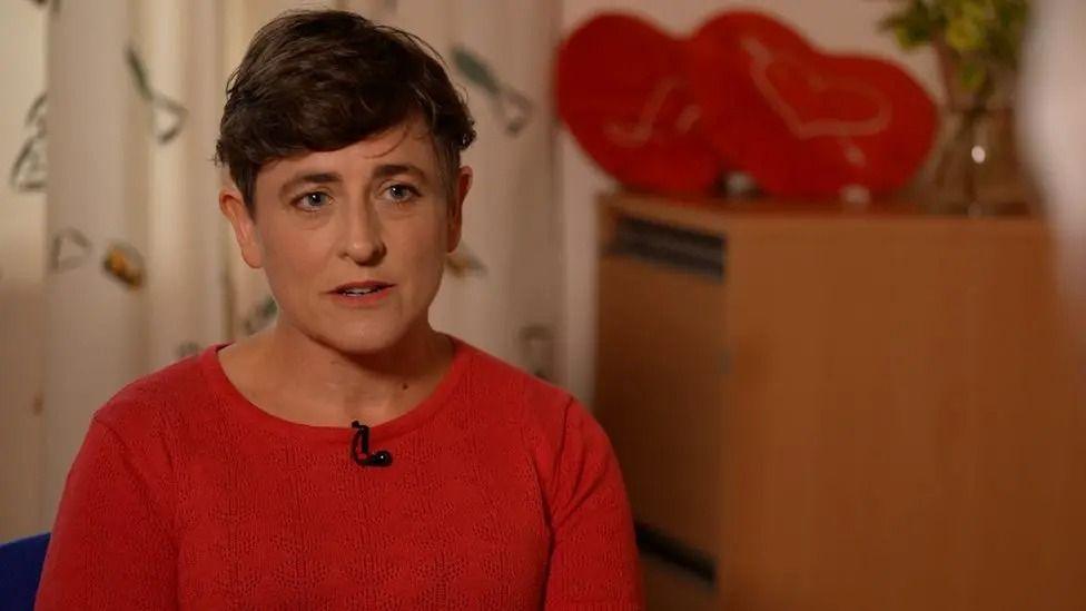 Helen Gittos is wearing a red jumper and sitting in front of some patterned curtains. She has dark brown short hair.