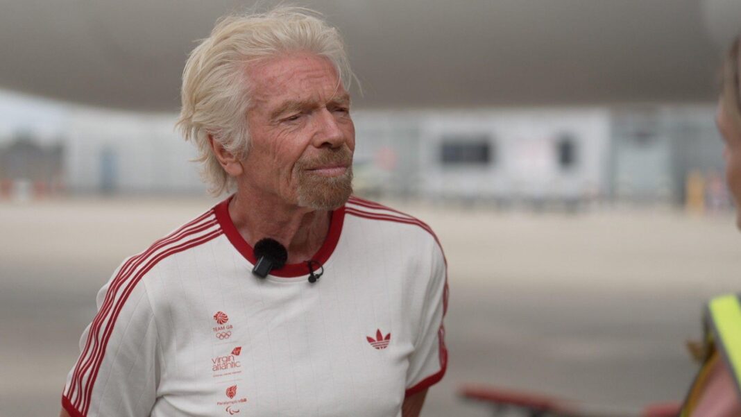 Flight price rises due to ‘foolish leaders’ starting wars, Virgin’s Branson says Richard Branson wearing a white t-shirt with the Team GB, Virgin Atlantic, and Paralympics GB logos in the right side of the chest with red lettering, an Adidas logo on the left side of the chest in red, and three red stripes on each arm. He is standing on the runway of Heathrow Airport. Behind him are a Virgin Atlantic plane and the terminal buildings in soft focus.