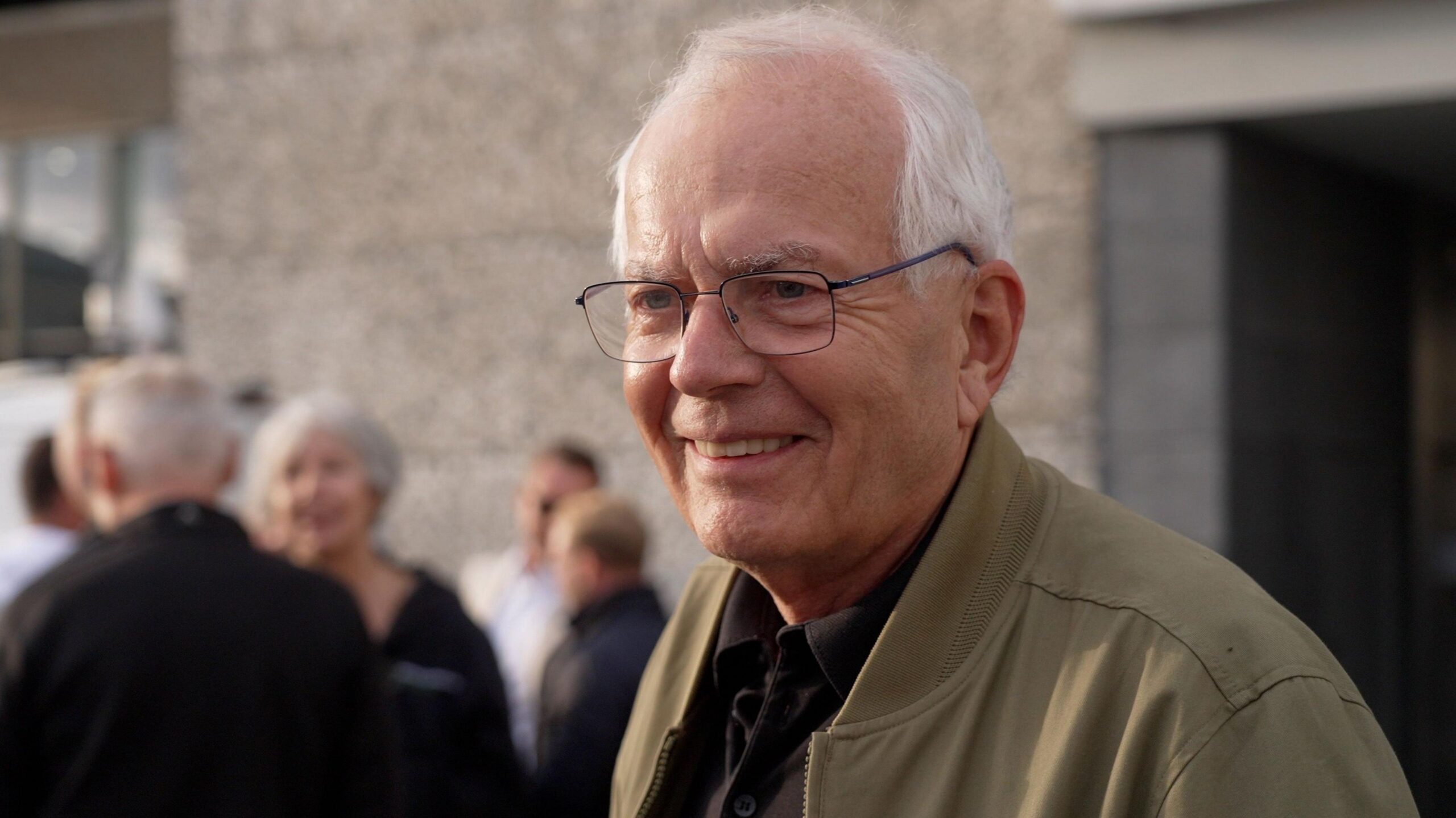 Reinhart. He is an older man with balding white hair. he has black rimmed glasses on and a beige unzipped jacket