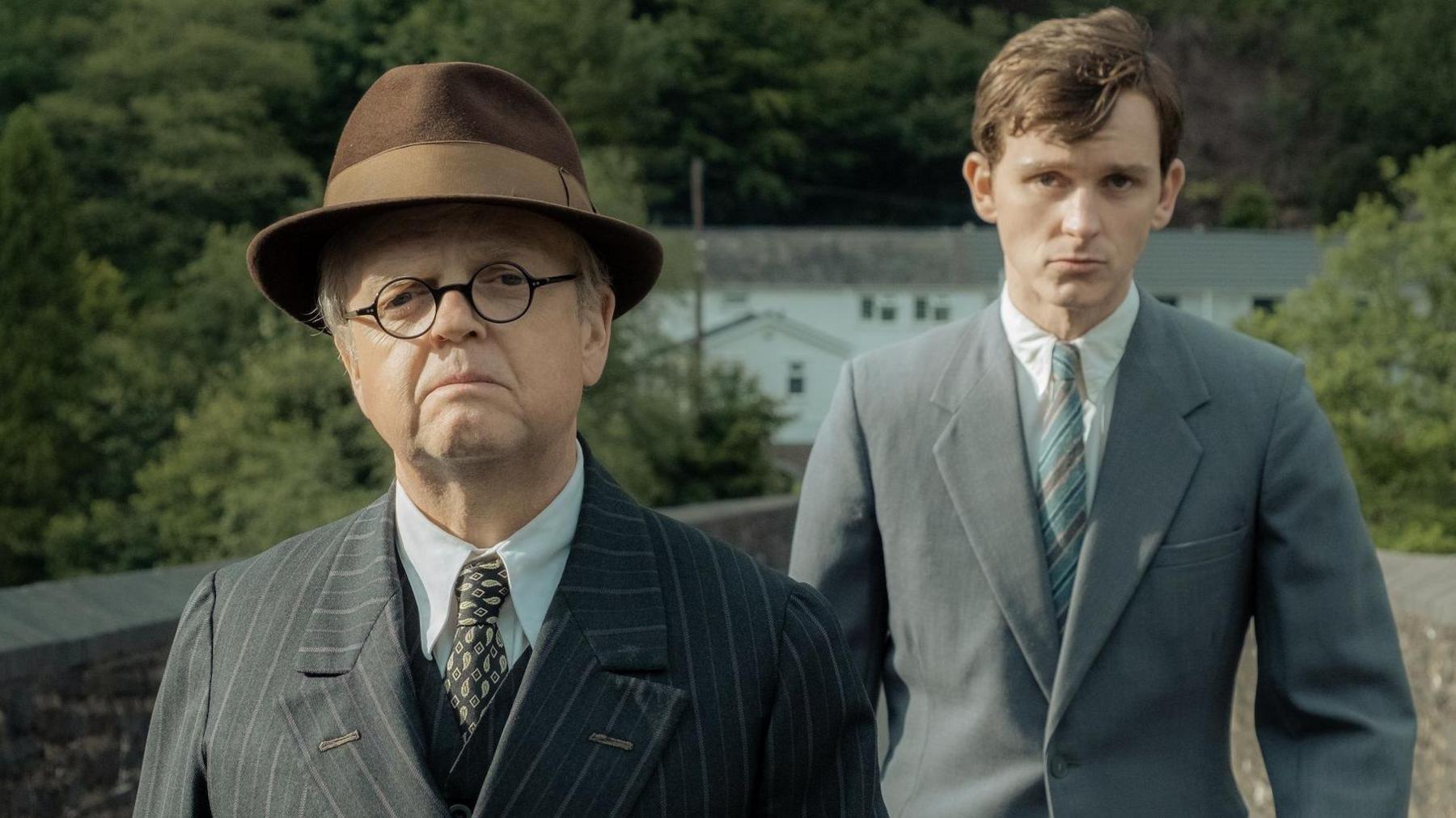 Toby Jones and Harry Lawtey in a scene in the film, walking across a footbridge with brick walls either side. They are both wearing a grey suit, white shirt and tie and Toby Jones, playing Philip Burton, wears glasses and walks ahead of Harry Lawtey