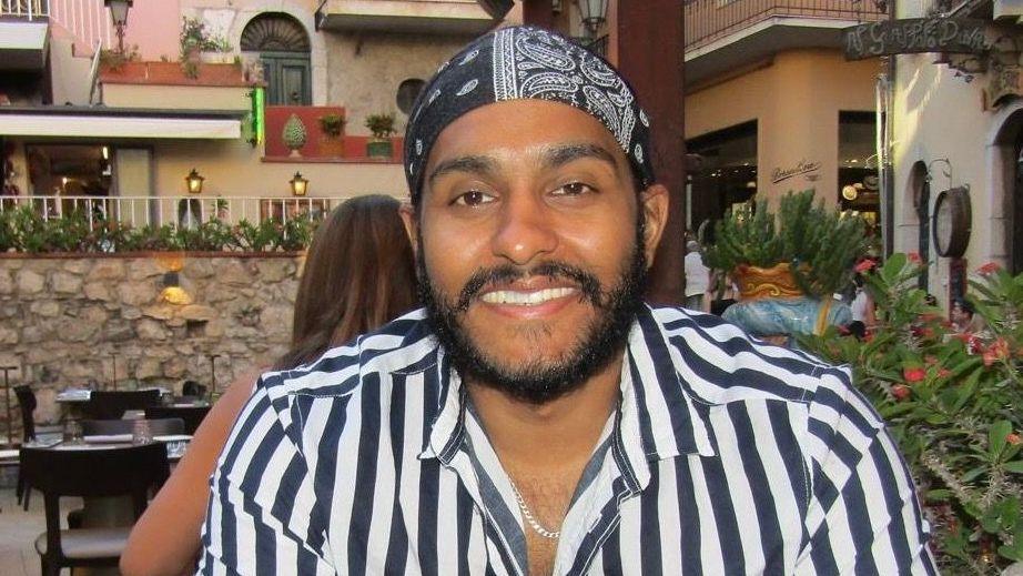 Amarpreet Singh sitting at an outdoor table smiling at the camera. He has a trim beard and moustache and is wearing a black and white patterned bandana on his head and a black and white striped shirt.