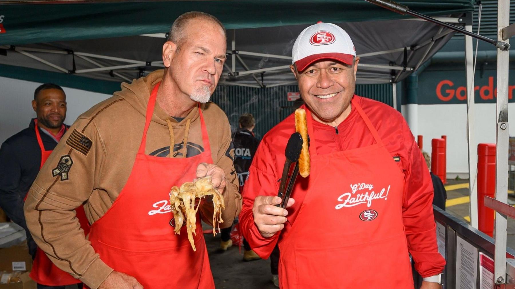 Two middle aged men in red aprons hold up a sausage and fried onions for the camera