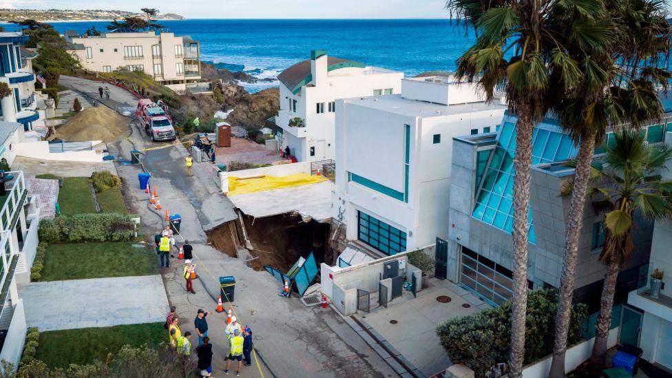 A large sinkhole is seen outside a mansion in Malibu, with emergency servicves attending, and palm trees and the ocean seen in the background