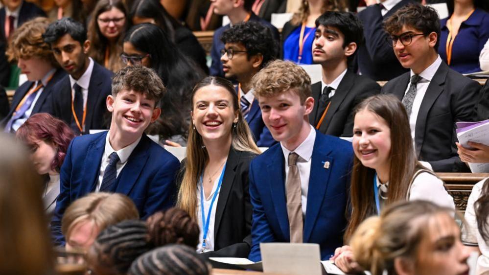 Members of Youth Parliament smile while sat on the benches of the House of Commons.