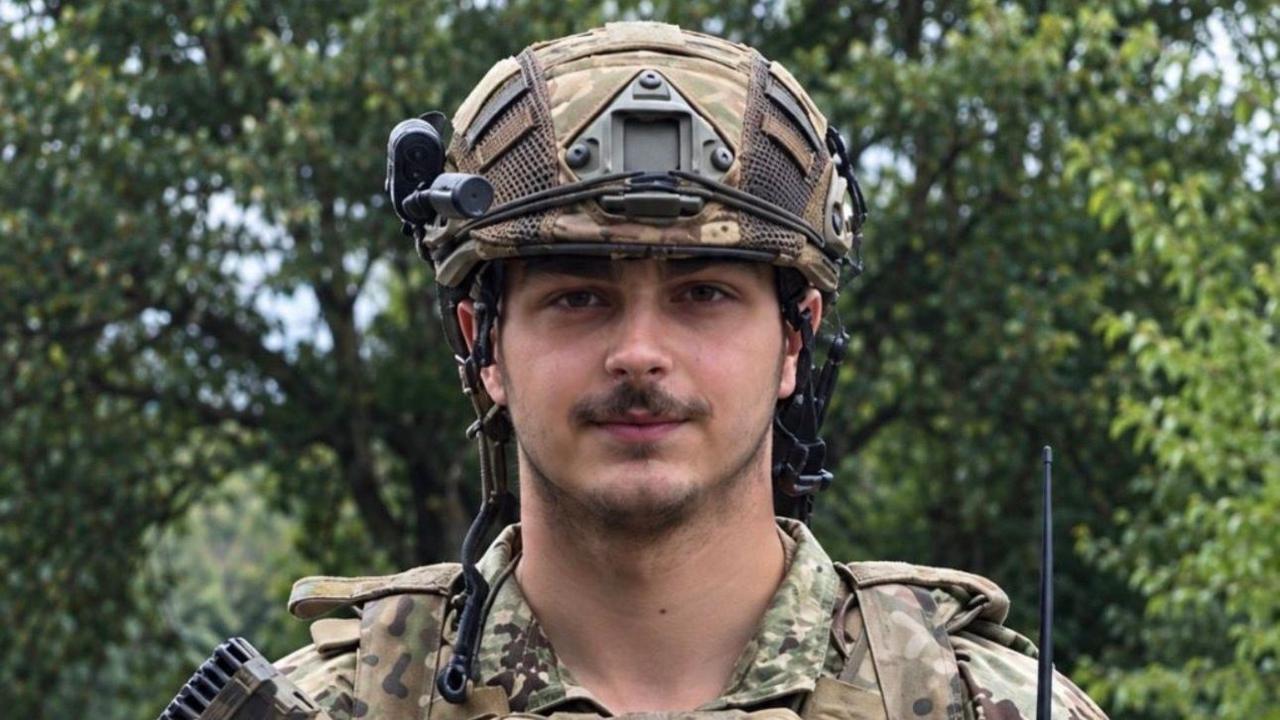 A young soldier with a moustache in a military helmet and army uniform with trees behind him