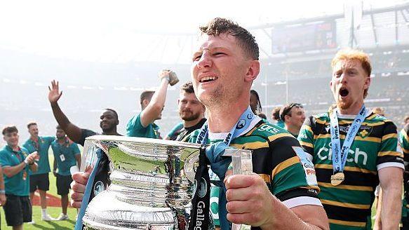 Fraser Dingwall holds the Prem trophy on the pitch at Twickenham after the 2026 Prem final. He is wearing a green, black and gold Northampton Saints jersey and his team-mates are celebrating in the background.