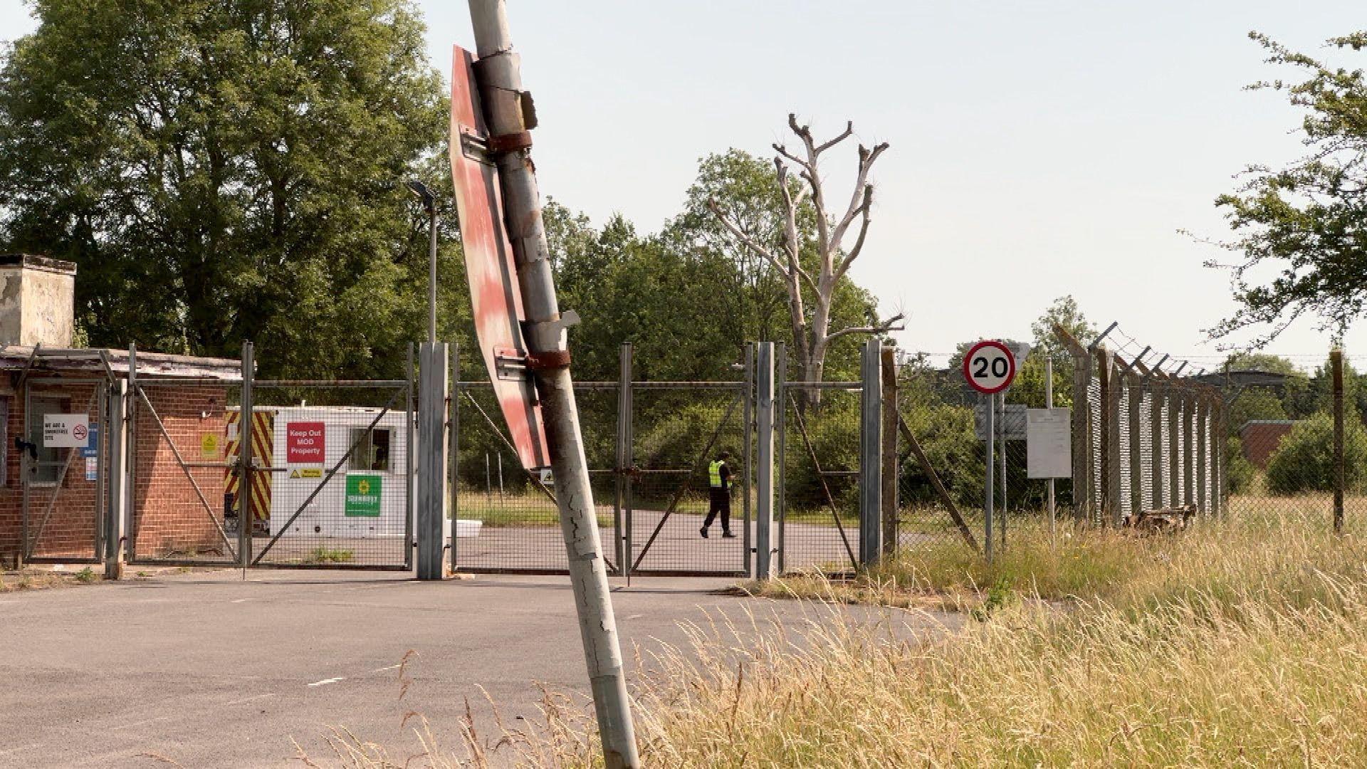 The gates outside MOD Bicester Site A. A security guard is patrolling behind gates and a barbed wire fence.