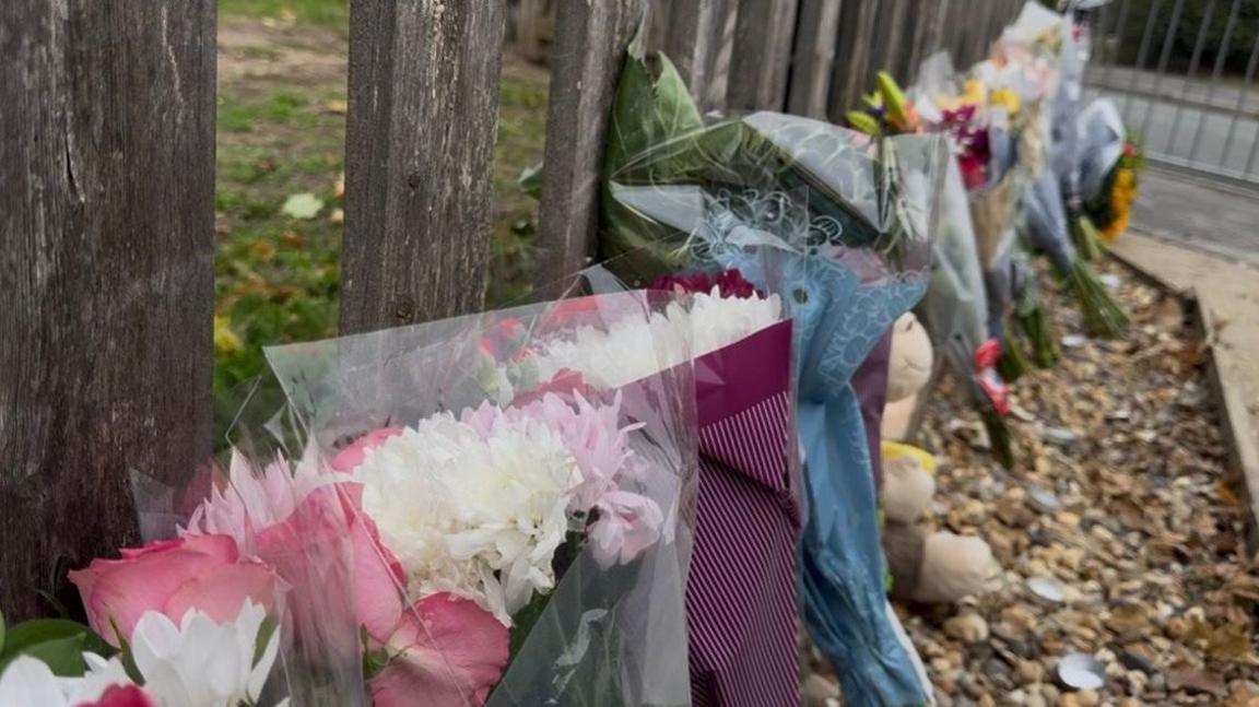 Bunches of flowers and soft toys are propped against a wooden fence.