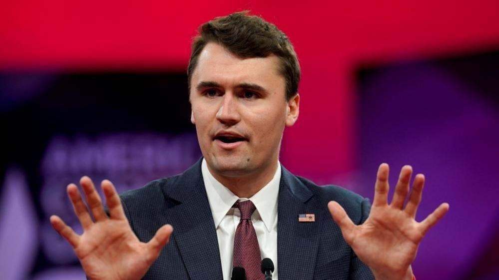 Charlie Kirk gestures with his hands as he talks in front of a red and purple backgroundi