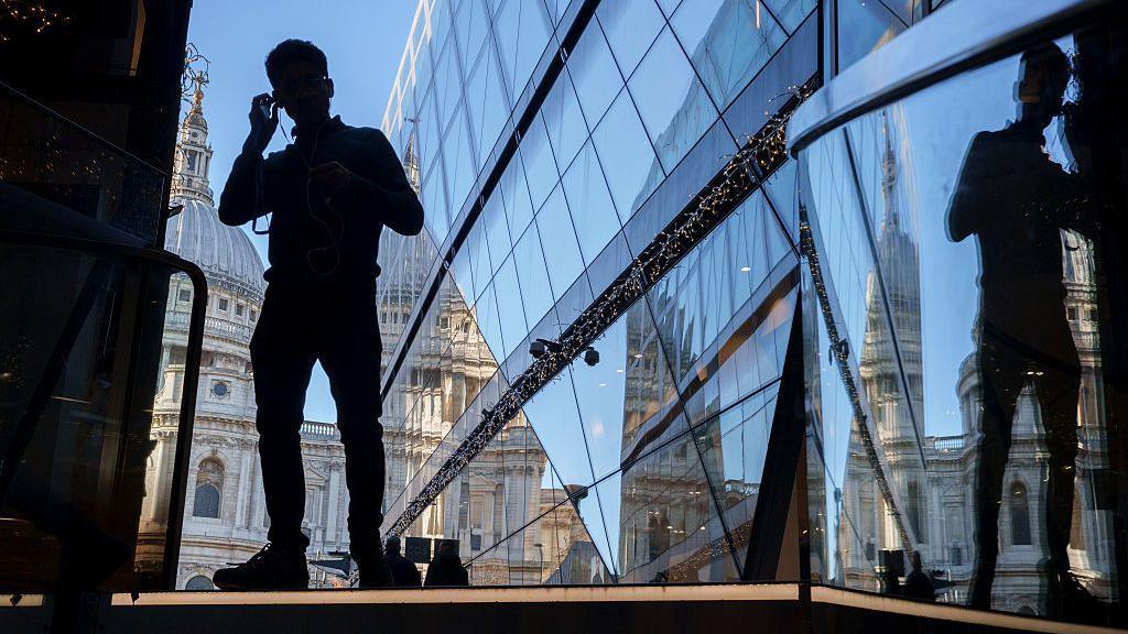 A silhouetted man puts headphones in his ears, standing next to a modern glass building reflecting St Paul's cathedral in the background.