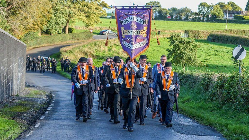 Orangemen walk down a road, behind them is a police presence