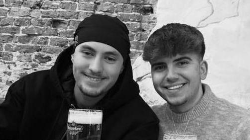 A black and white photo of Eddie and Jayden, smiling side by side. They are both holding pints or beer. Jayden has a hat on and has short stubble and is wearing a black hoodie. Eddie has short dark hair and is wearing a jumper.