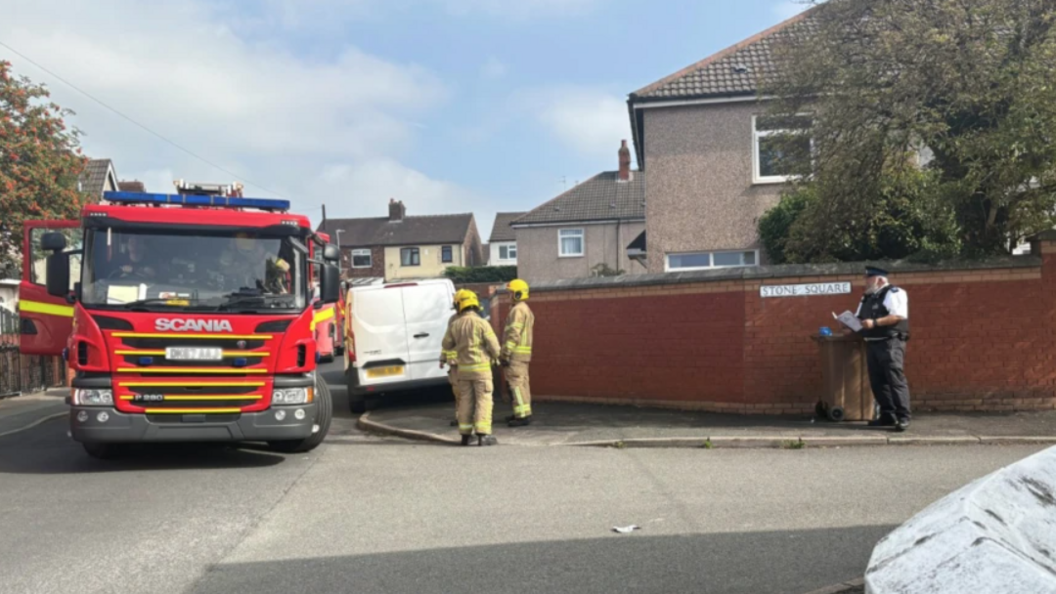 A fire engine is parked on a road outside a row of houses. A police officer and two firefighters are standing close by.