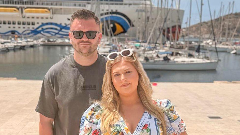 Conroy and Amber stand in front of a cruise ship docked in a harbour. It's a sunny day; he wears a black T-shirt and sunglasses. She wears a flowery short-sleeved shirt.