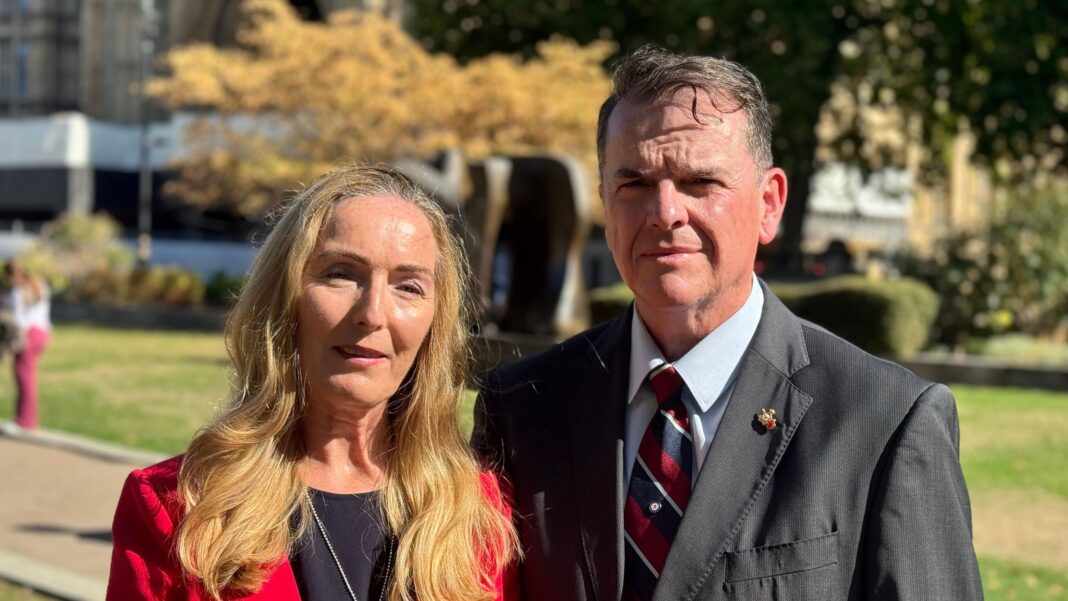 A portrait shot of Paula McGowan and her husband Tom outside parliament. She is wearing a red dress and has long blonde hair. He is wearing a suit and a blue shirt.