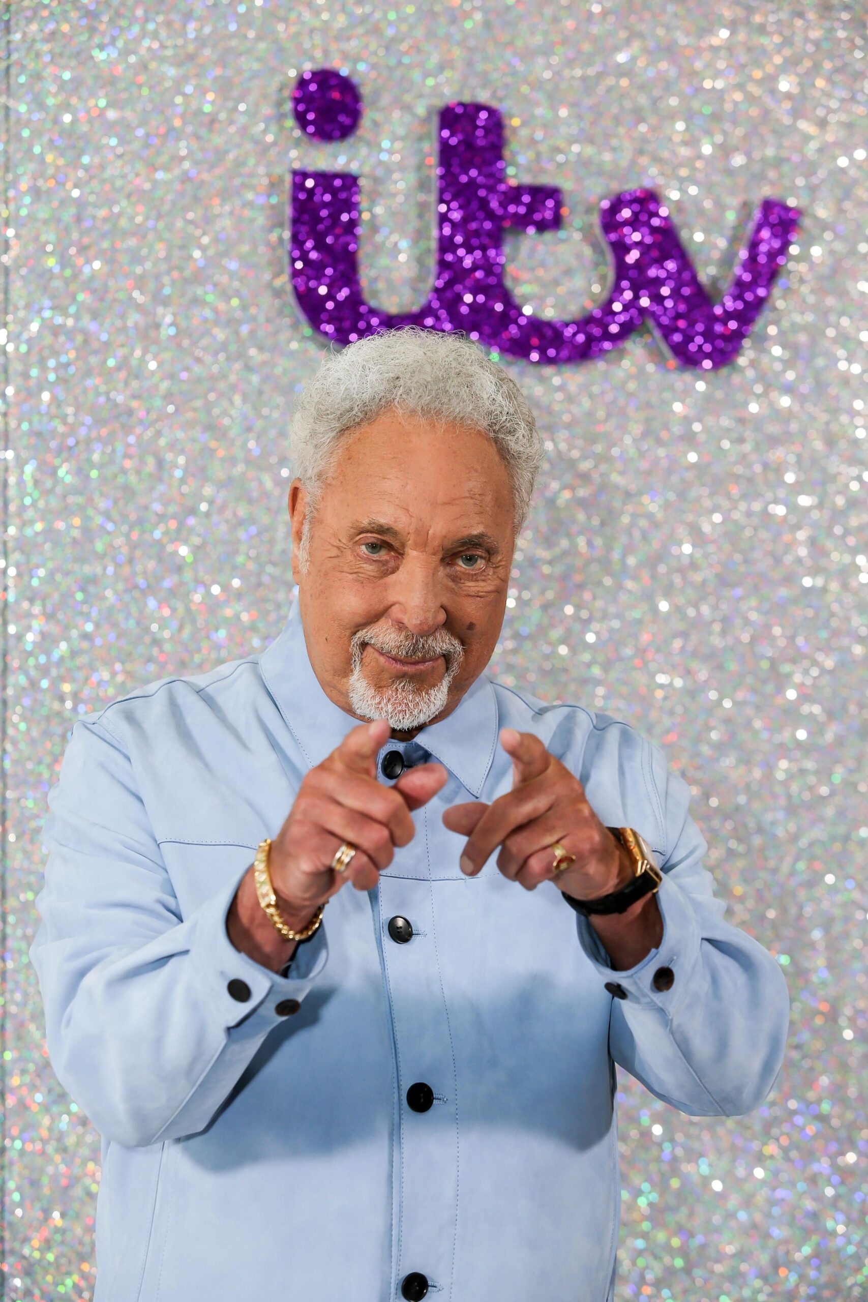 Sir Tom Jones poses pointing both index fingers at the camera, he is stood in front of a sparkly white backdrop with ITV written in sparkly purple letters. Sir Tom has white hair and is wearing a pale blue shirt.